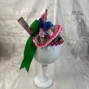 May include: A white milk glass goblet topped with a decorative arrangement featuring a bright green ribbon, a blue ruffled fabric accent, and a beige fabric piece with colorful polka dots and a pink pom-pom trim, plus a small rectangular card.