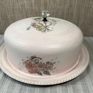 May include: A pale pink cake dome and matching plate featuring a clear crystal knob handle. The dome is decorated with a floral transfer on the side and a clockwork gear design on the top, both rendered in muted earth tones and black ink.