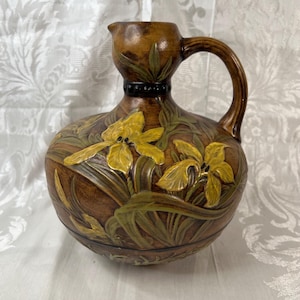 May include: A brown ceramic pitcher featuring a relief design of yellow iris flowers and green leaves. The vessel has a rounded body, a narrow neck with a dark band, and a curved handle extending from the neck to the shoulder of the piece.