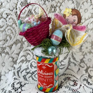 May include: A decorative arrangement featuring a red tin labeled Delicious After Dinner Mints, topped with a pink woven basket containing a small rabbit and eggs, a paper doll figure with a yellow flower, and green moss accents.