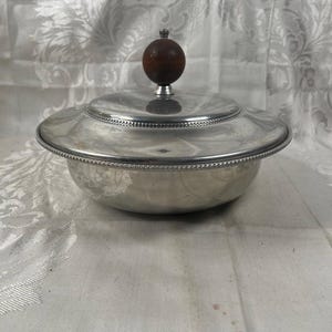 May include: A round silver-toned metal serving bowl with a matching lid. The lid features a decorative beaded edge and a dark brown spherical wooden knob handle centered on top. The bowl has a smooth, polished finish and a wide, flat rim.