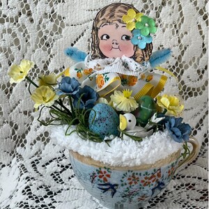 May include: A decorative teacup filled with white faux fur, featuring a vintage-style paper doll head with floral hair accents, blue pipe cleaner wings, yellow and blue fabric flowers, a speckled blue egg, and a small white bird figurine.