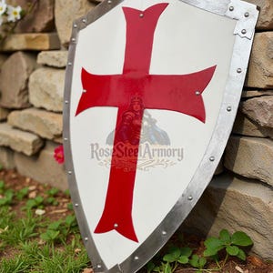 May include: A white shield with a large red cross design, framed by a silver metal border. The shield has a logo that reads "Rose Steel Armory". The shield is leaning against a stone wall.