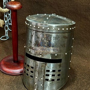 May include: A distressed steel helmet with a rectangular face opening and small square eye holes. The helmet has a cylindrical shape and is secured with small, round rivets. The text "Rose Steel Armory" is visible on the side.