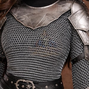 May include: A close-up of a medieval-style costume. The costume features a chainmail shirt, metal shoulder armor, and a black leather belt with a decorative buckle. The armor is silver-toned, and the belt has silver studs.