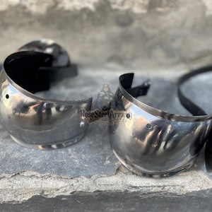 May include: Two polished steel arm guards, likely for historical reenactment or display. The curved pieces have a reflective surface and are connected by black straps. Each guard has small rivet details and a rounded shape.