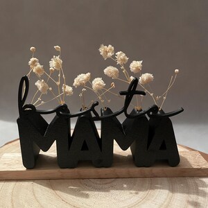 Könnte beinhalten: Schwarzes Metall-Blumenarrangement mit dem Wort "Mama" und dem Wort "Best" darüber. Das Arrangement enthält kleine weiße Blumen. Das Stück wird auf einer Holzoberfläche präsentiert.