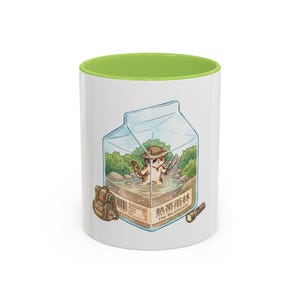 May include: White ceramic mug with a green interior. The mug features a cartoon illustration of a cat in a rainforest scene inside a milk carton. A backpack and flashlight are also depicted. The text "The Rainforest" is visible.