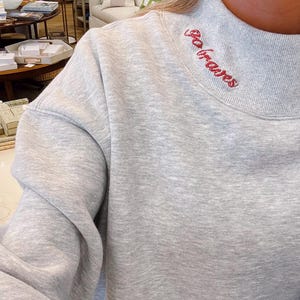 Puede incluir: Una sudadera gris claro con cuello alto. Las palabras "Go Braves" están bordadas en rojo en el cuello. La sudadera tiene mangas largas y un corte holgado, adecuada para uso casual.