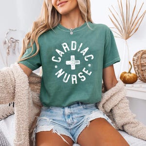 Könnte beinhalten: Ein grünes T-Shirt mit dem weißen Schriftzug "CARDIAC NURSE" in einem kreisförmigen Design mit einem Kreuz in der Mitte. Das Shirt hat kurze Ärmel und ist aus weichem Material. Das Model trägt Jeansshorts und eine cremefarbene Strickjacke.