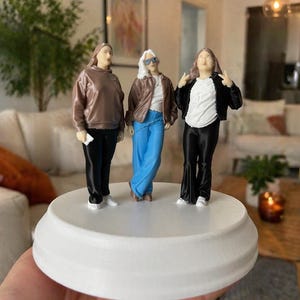 May include: A 3D-printed figurine of three figures on a white circular base. The figures are dressed in diverse outfits: a brown top and black trousers, a brown jacket and blue jeans, and a black jacket and black trousers. The base is held in a hand.