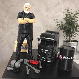 Puede incluir: Una figura impresa en 3D de una persona con barba blanca, camisa y pantalones negros, junto a un modelo de camión MAN negro, un barril negro con la etiqueta Trailer Service, una caja de madera y varias herramientas en miniatura como llaves.