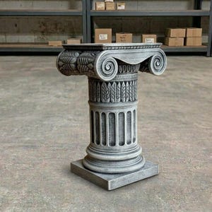 May include: A gray, decorative pedestal with an ornate, classical design. The pedestal features a square base, a fluted column, and a detailed capital with scroll and leaf patterns. The overall height is approximately 30 inches.