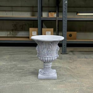 May include: A light gray stone urn planter featuring a wide, flared rim and a body decorated with vertical acanthus leaf carvings. The vessel rests on a fluted pedestal base that sits atop a square plinth, showcasing a classic, ornate design.