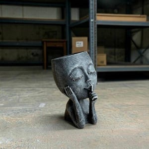 May include: A dark gray, textured planter shaped like a human head with closed eyes and long eyelashes. The figure has one hand resting against its cheek and the other hand held up to its mouth in a shushing gesture, creating a hollow vessel top.