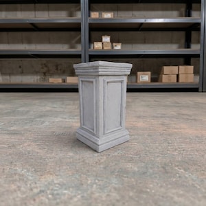 May include: A light gray rectangular pedestal with a flat top, a recessed panel design on the front face, and a stepped base. The structure features clean, straight lines and a smooth, uniform finish, standing upright as a display stand.