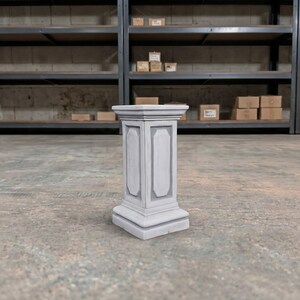 May include: A light gray rectangular pedestal featuring a stepped base and a matching stepped top. The central column of the pedestal is decorated with recessed vertical panels on each side, creating a structured and symmetrical appearance.