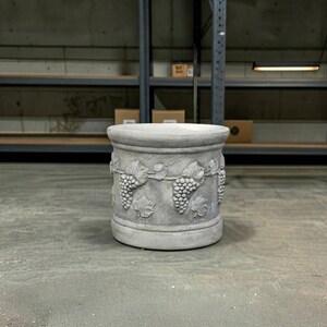 May include: A cylindrical gray stone planter featuring a relief pattern of grape clusters and vine leaves encircling the exterior. The object has a smooth, flat top rim and a solid base, with a weathered, textured finish throughout the surface.