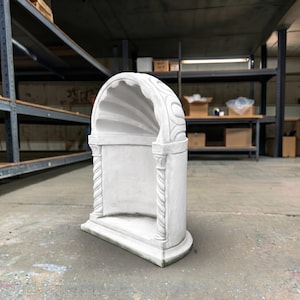 May include: A white, arched stone niche featuring a scalloped interior ceiling and two spiraled columns on the front sides. The structure has a flat base and a curved top, with decorative carved details on the exterior frame and column capitals.