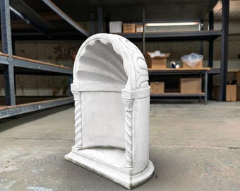May include: A white, arched stone niche featuring a scalloped interior ceiling and two spiraled columns on the front sides. The structure has a flat base and a curved top, with decorative carved details on the exterior frame and column capitals.