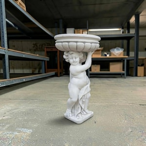 May include: A white stone-colored statue of a cherub standing on a rectangular base. The cherub is depicted with curly hair, holding a large, round, fluted basin above its head with both arms. A piece of draped fabric covers the lower body.