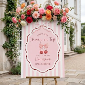 May include: A baby shower welcome sign with pink and white stripes, featuring the text "She's the Cherry on Top" and "Welcome to Vanessa's Baby Shower." The sign is adorned with a cherry illustration and a floral arrangement of pink, red, and orange roses.
