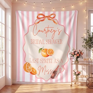 May include: A backdrop with pink and white stripes, featuring the text "Courtney's Bridal Shower" and "Last Spritz as a Miss". The design includes orange illustrations and a bow.