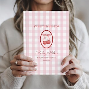 Op de afbeelding: Een gastenboek voor een babyshower met een roze en witte ruitjespatroon. De omslag toont de tekst "Sweet Wishes For Baby Mika" en een kersenillustratie. Het boek is opengehouden en toont de titel en de datum augustus 2026.