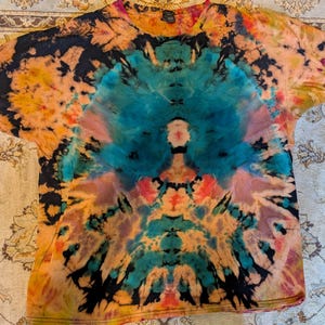 Può includere: Una t-shirt a maniche corte con un vivace motivo tie-dye caratterizzato da un'esplosione centrale di color ottanio circondata da vortici neri, arancioni, gialli e rosa. Il design è simmetrico e si irradia dal centro verso le maniche e l'orlo.