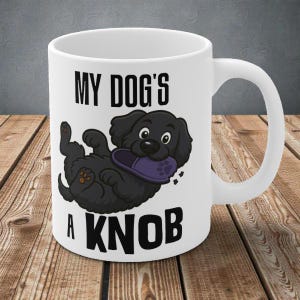 Puede incluir: Taza de cerámica blanca con una ilustración de un perro negro de dibujos animados sosteniendo una zapatilla morada. El texto "MY DOG'S A KNOB" está impreso. La taza tiene un asa blanca y está sobre una superficie de madera.
