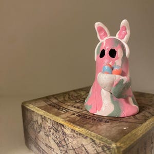 Può includere: Statuina in argilla fatta a mano di un fantasma con orecchie da coniglio, che tiene un cestino di uova di Pasqua. Il fantasma è rosa, bianco e verde con occhi neri. La statuina è su una scatola decorativa con un disegno di mappa del mondo.