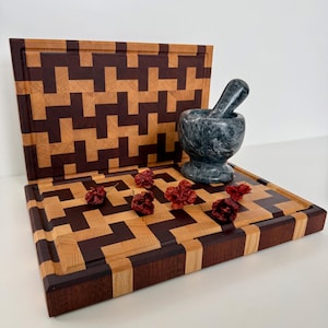 Può includere: Due taglieri in legno con un motivo geometrico marrone chiaro e scuro. Un mortaio e un pestello grigio scuro sono su una tavola, con diversi peperoncini rossi secchi. Le tavole hanno un incavo per il succo.