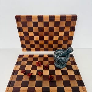 Può includere: Due taglieri in legno con un motivo a scacchiera nei toni marrone chiaro e scuro. Un mortaio e un pestello grigi e peperoncini rossi secchi sono su una tavola. Le tavole hanno una scanalatura per il succo sui bordi.