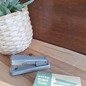 May include: A vintage gray stapler and a box of staples sit on a wooden surface. The stapler is metallic gray and has a classic design. The box of staples is light blue and white, with the text "BATES Standard Staples". A woven basket with a green plant is in the background.