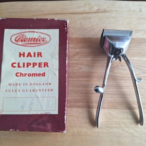May include: A vintage Premier Hair Clipper, chromed, alongside its original maroon box. The box displays the Premier logo and text: "Hair Clipper Chromed Made in England Fully Guaranteed". The manual hair clipper is made of shiny metal.