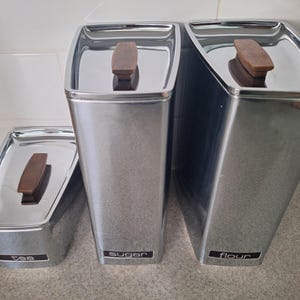 May include: A trio of stainless steel kitchen canisters with wooden handles. The canisters are labelled "tea", "sugar", and "flour" in a black font. The lids are flat and the canisters have a rectangular shape.