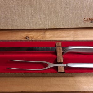 May include: A carving set with a serrated blade knife and a two-pronged fork, presented in a red-lined box. The knife and fork have silver-colored handles. The box is tan with the "GERBER" logo.