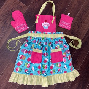 May include: A pink and light blue apron with a cupcake pattern, two pink pockets, and a ruffled hem, accompanied by a matching pink oven mitt and a square pot holder. Both the apron bib and the accessories feature the text Olivia's kitchen.