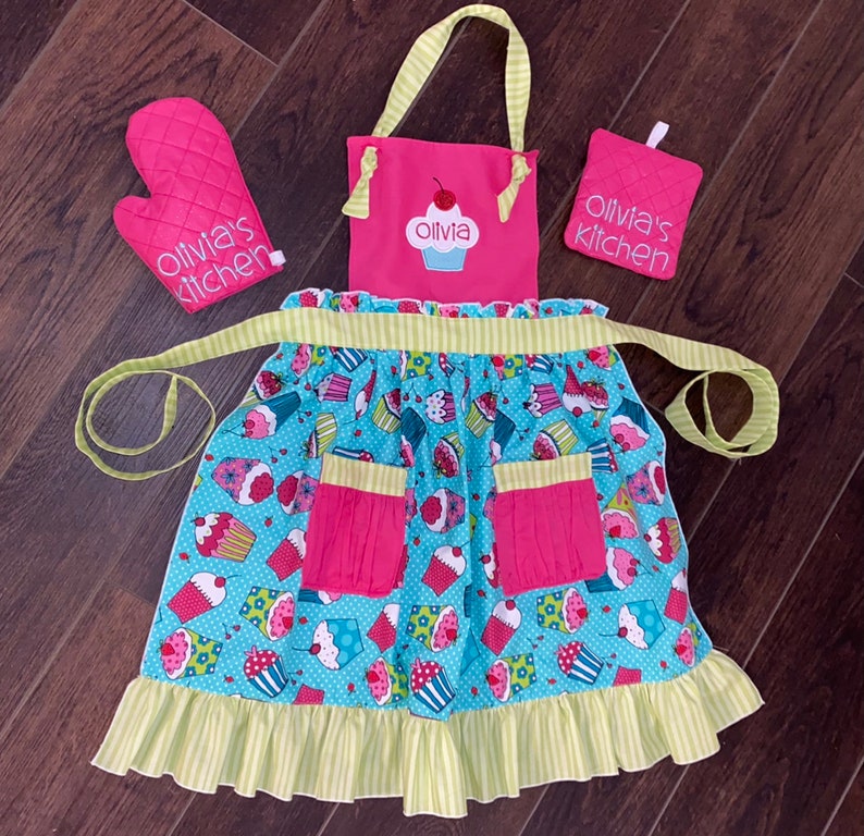 May include: A pink and light blue apron with a cupcake pattern, two pink pockets, and a ruffled hem, accompanied by a matching pink oven mitt and a square pot holder. Both the apron bib and the accessories feature the text Olivia's kitchen.
