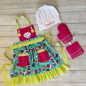 May include: A kitchen set featuring a teal apron with a cupcake pattern, a pink bib with the name Emersyn, a white chef hat embroidered with Emersyn, and a pink quilted oven mitt and pot holder both embroidered with the text Emersyn's kitchen.