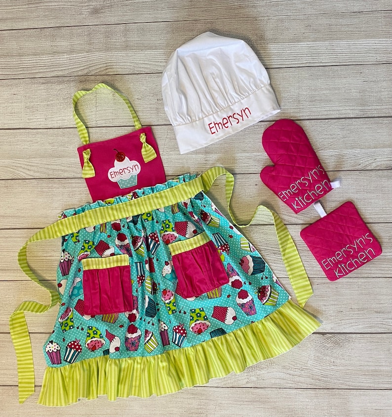 May include: A kitchen set featuring a teal apron with a cupcake pattern, a pink bib with the name Emersyn, a white chef hat embroidered with Emersyn, and a pink quilted oven mitt and pot holder both embroidered with the text Emersyn's kitchen.