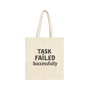 Puede incluir: Un bolso tote de color crema con texto negro que dice "TASK FAILED Successfully". El bolso tiene dos asas y está hecho de un material similar a la lona. El texto está centrado en la parte delantera del bolso.