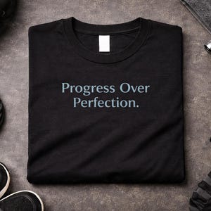 Pode incluir: Camiseta preta com o texto "Progress Over Perfection" em azul claro. A camisa é cercada por equipamentos de fitness, incluindo uma corda de pular, halteres e tênis esportivos, tudo em uma superfície cinza.