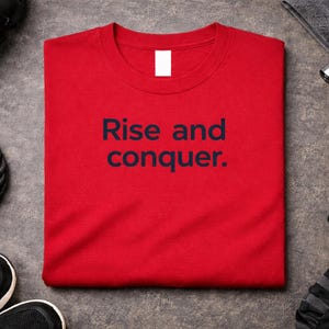 Pode incluir: Uma camiseta vermelha com o texto azul marinho "Rise and conquer". A imagem inclui equipamentos de fitness, como uma garrafa de água preta, uma corda, halteres e tênis pretos.