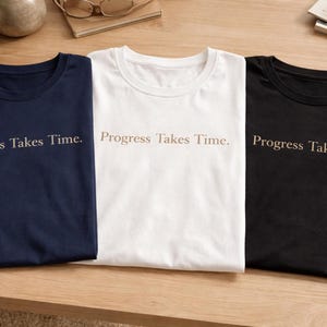 Pode incluir: Três camisetas dobradas em azul marinho, branco e preto, cada uma com a frase "Progress Takes Time" impressa em marrom claro. As camisetas estão dispostas sobre uma superfície de madeira clara.