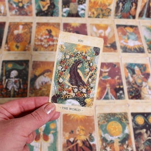 Peut inclure: Une carte de tarot d'un jeu, tenue dans une main, représentant une illustration d'une femme entourée d'une couronne florale. La carte est intitulée "XXI - THE WORLD" et est placée sur un fond d'autres cartes de tarot.