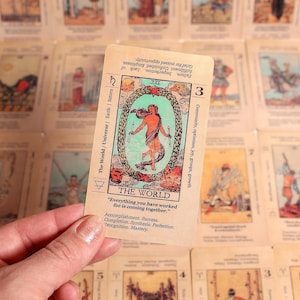 Könnte beinhalten: Eine Tarotkarte aus einem Deck, die eine Person in einem Lorbeerkranz zeigt, mit der Aufschrift "DIE WELT". Die Karte wird über einem Hintergrund aus anderen Tarotkarten gehalten. Das Design der Karte enthält Text und Symbole.