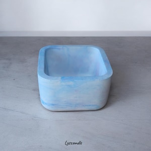 Peut inclure: Un récipient carré en béton bleu clair marbré. Le récipient a des coins arrondis et une finition lisse. L'intérieur est d'une nuance de bleu plus claire. Le mot "Lyscandle" est imprimé en bas.