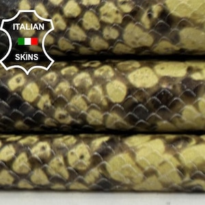 Puede incluir: Primer plano de pieles de cuero apiladas y texturizadas con un patrón de piel de serpiente. Las pieles presentan una mezcla de escamas negras, marrones y amarillas. La imagen incluye el logotipo "Italian Skins".