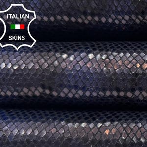 May include: Three rolls of navy blue leather with a scale-like texture are shown in close-up. The leather has a subtle sheen and reflective accents. The Italian Skins logo is visible. The rolls are tightly wound.